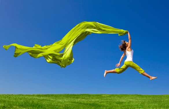 Beautiful young woman jumping on a green meadow with a colored tissue